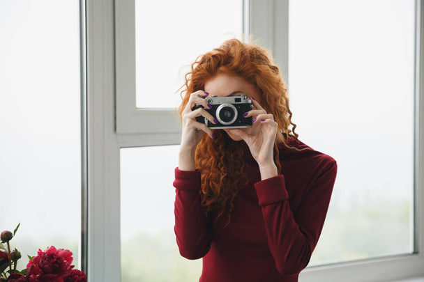 Redhead lady indoors near flowers holding camera. - Foto, afbeelding