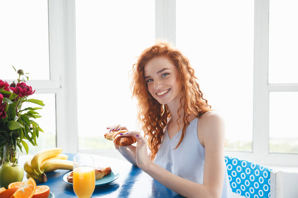 Happy young redhead lady sitting at the table eating croissant - Fotoğraf, Görsel