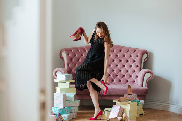 Happy young lady standing near sofa indoors choosing shoes - Photo, image