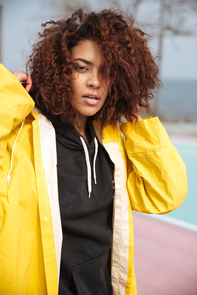 Concentrated african curly young woman wearing yellow coat - Фото, изображение