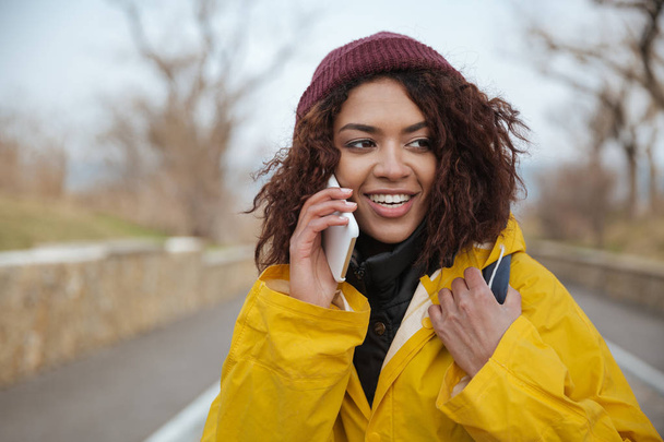 Happy african young lady wearing yellow coat talking by phone. - Photo, image