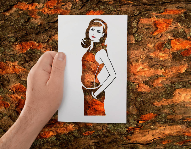 Egy kéz tartja a papír rajzsablon egy fiatal hosszú hajú lány a háttérben a fa kéreg. - Fotó, kép