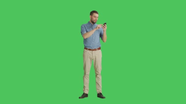 Long Shot of a Handsome Man Holding Smartphone with One Hand and Making Swiping Touching Gestures with Another. Tablet and Background are Green Screen. - Felvétel, videó