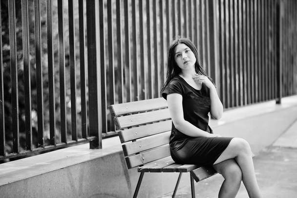 Brunette girl at black dress sitting on bench and posing at stre - Fotó, kép