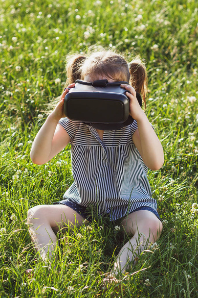 Little girl plays game with virtual reality glasses outdoors - Φωτογραφία, εικόνα