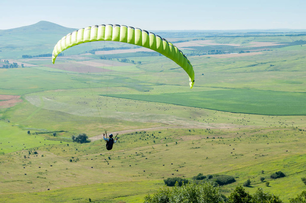 Paragliding pilot flying fields against the blue sky and mountain landscape - Фото, изображение