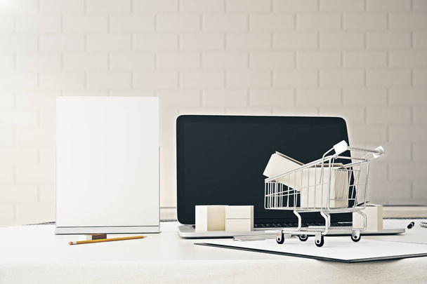 Close up of office desktop with empty laptop screen, blank poster and tiny shopping cart. Ad concept. - Fotó, kép