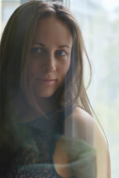 Вистрілив через скло. Крупним планом портрет щасливі кавказьких жінка з блакитними очима, посміхаючись і, дивлячись на камеру позує перед фону вікно. - Фото, зображення