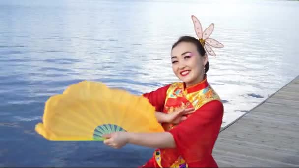 όμορφη κοπέλα κινέζικα σε παραδοσιακά κόκκινο φόρεμα κινέζικο με κίτρινο οπαδός στις όχθες του ποταμού - Πλάνα, βίντεο