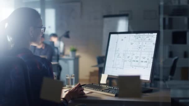 Tarde en la noche en la Oficina de Ingeniería Diseñadora femenina trabaja en una computadora personal que muestra planos de su proyecto. En el fondo, sus colegas trabajando
. - Imágenes, Vídeo