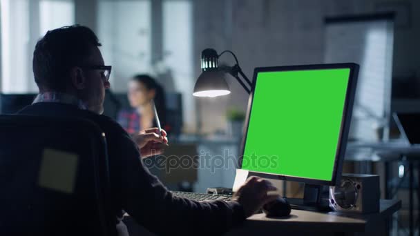 Tarde en la noche Ingeniero Industrial se sienta en su escritorio trabajando en una computadora con pantalla verde. La oficina es moderna. En el fondo Colegas están trabajando en sus escritorios
. - Imágenes, Vídeo
