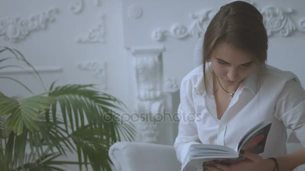 Close up portrait of attractive young woman reading a book while relaxing in chair in the living room. - Video