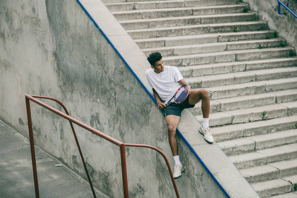 An athlete with a tennis racket sits on a concrete railing of steps. - Foto, Imagem