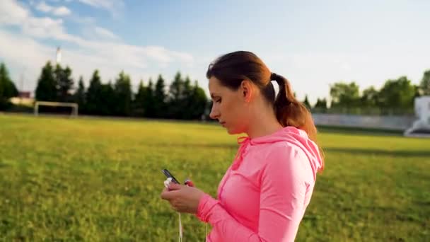 Woman inserts headphones into ears, preparing for a run at the stadium - Felvétel, videó
