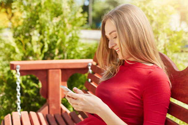 Sideways portrait of teenage girl with light hair sitting at bench in park using smartphone smiling while recieving message from her boyfriend. Attractive woman surfing internet using cell phone - Zdjęcie, obraz