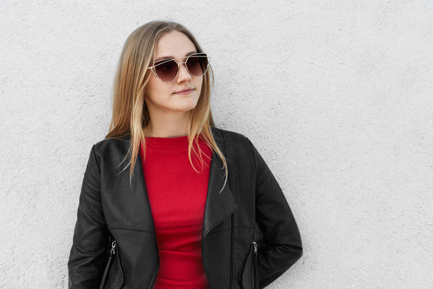Sexy blonde female in sunglasses and black leather coat standing against white concrete wall with blank space for your advertisment looking aside having thoughtful expression and pleasant look - Photo, image