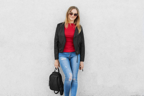 Fashionable woman dressed in red sweater, jeans and black leather jacket wearing trendy sunglasses holding black backpack in her hand posing against white background. Fashion and beauty concept - Fotoğraf, Görsel