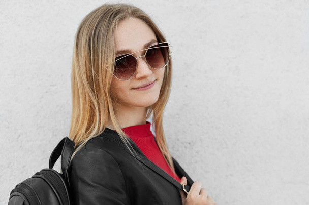 Sideways portrait of stylish teenage girl with fair hair wearing big trendy sunglasses, leather jacket and holding black backpack isolated over white background. People, fashion and youth concept - Φωτογραφία, εικόνα