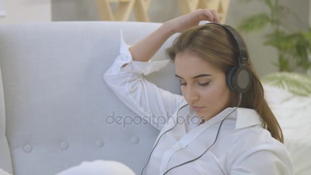 Portrait of young woman in casual shirt listening music in headphones while relaxing on the chair in home. - Filmagem, Vídeo