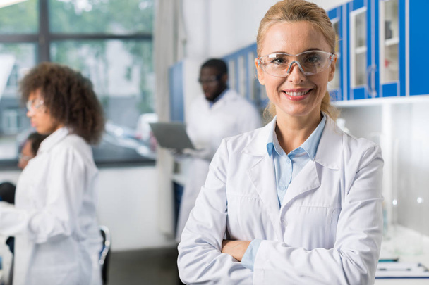Portret uśmiechający się kobiety w biały płaszcz i okulary ochronne w nowoczesnym laboratorium, Kobieta naukowiec przez zespół naukowców zajęty - Zdjęcie, obraz