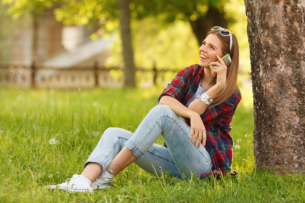 Genç mutlu kadın yaz Şehir Parkı çimlere oturup cep telefonuyla konuşuyor. Bir akıllı telefon, açık ile güzel modern kız - Fotoğraf, Görsel