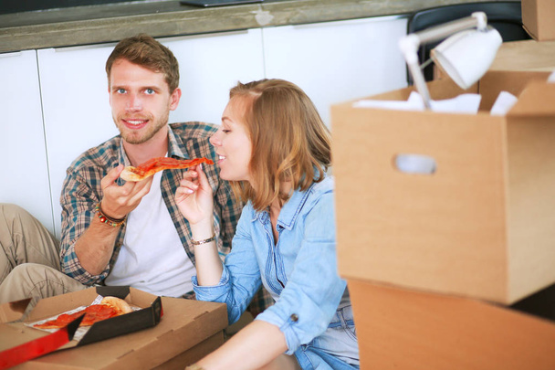 Young couple have a pizza lunch break on the floor after moving into a new home with boxes around them - Foto, Bild