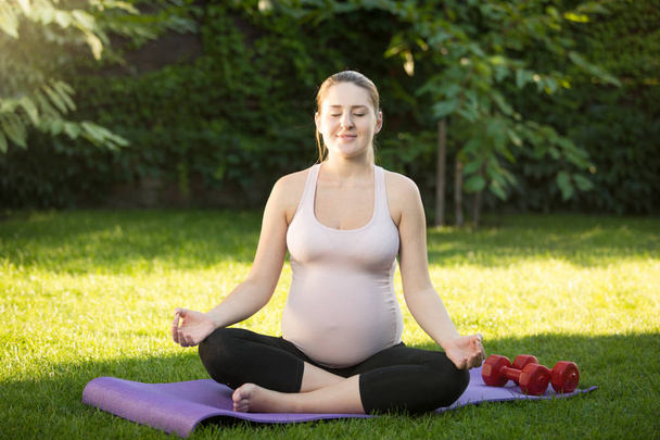 Όμορφη χαμογελαστή έγκυος γυναίκα κάθεται στο λωτό παρουσιάζουν στο γρασίδι  - Φωτογραφία, εικόνα