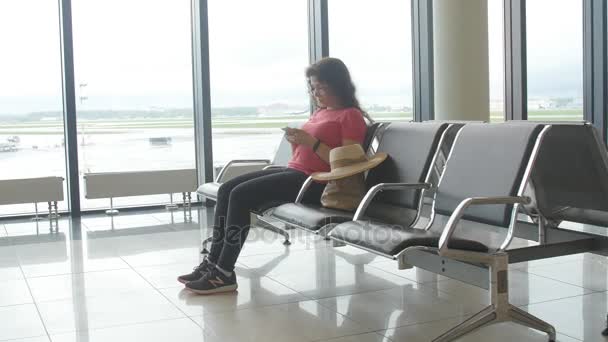 eine junge Frau am Flughafen, die auf ihre Abreise wartet und auf ihr Smartphone schaut - Filmmaterial, Video