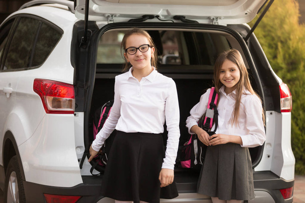 Muotokuva kahdesta hymyilevästä tytöstä ottamassa koululaukkuja ulos autosta
 - Valokuva, kuva