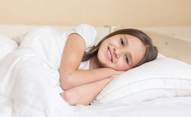 Χαριτωμένο χαμογελαστό κορίτσι ξαπλωμένο στο μαξιλάρι και βλέπει τα φωτογραφικών μηχανών - Φωτογραφία, εικόνα