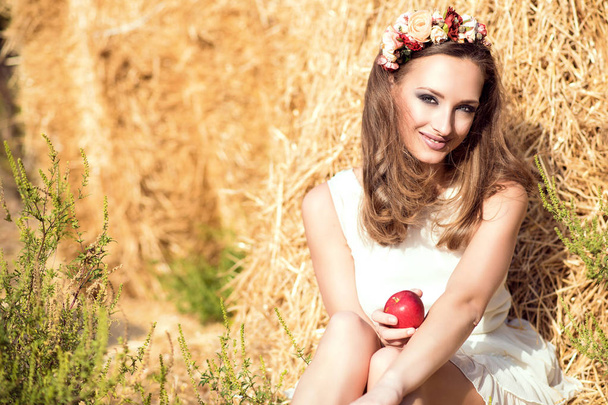 Портрет красивой улыбающейся девушки в белом летнем платье и цветочном венке на голове, сидящей у стога сена и держащей в руке красное яблоко
 - Фото, изображение