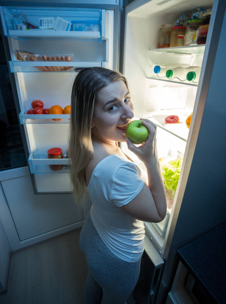 Portrait of hungry woman biting apple at night on kitchen - Photo, Image