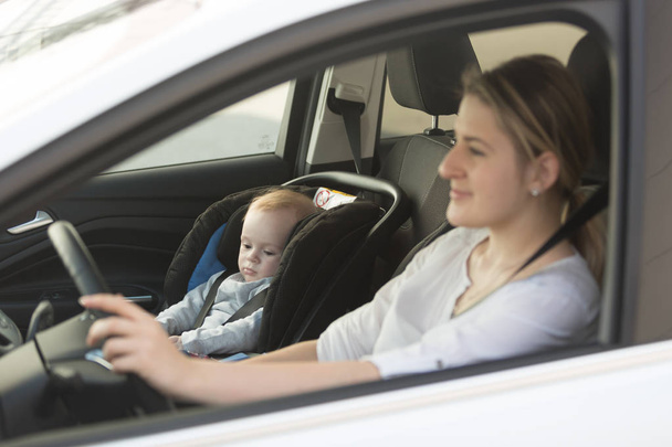 Young mother driving car with her baby boy sitting in car in bab - 写真・画像