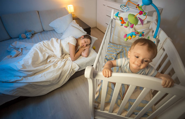 Jovem mãe cansada adormeceu ao lado do berço do bebê à noite
 - Foto, Imagem