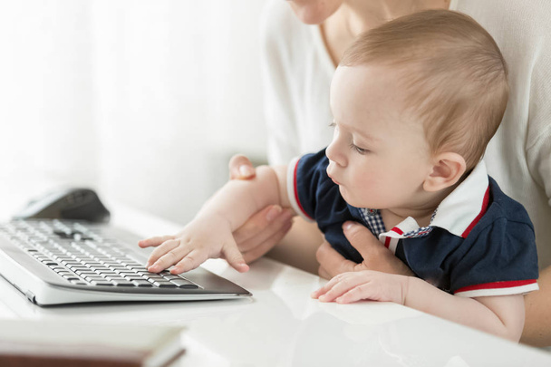 Νέοι η μητέρα κάθεται στον υπολογιστή και κρατώντας το μωρό στην αγκαλιά - Φωτογραφία, εικόνα