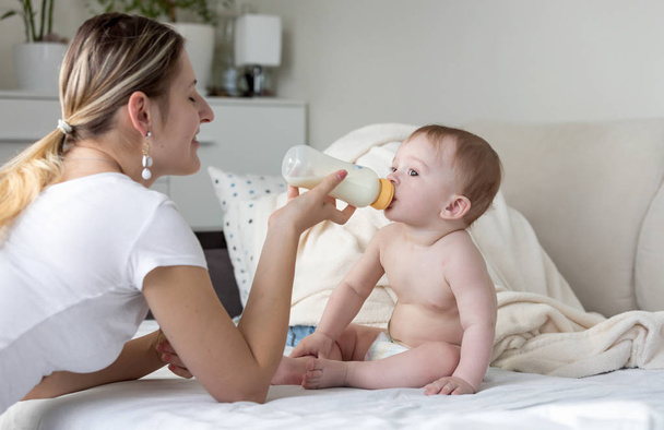 Yatağın üstüne oturan ve şişe süt beslenme bebek çocuk portresi - Fotoğraf, Görsel