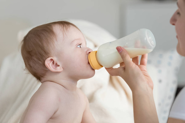 Mother giving milk from bottle to her baby son sitting on sofa - Photo, Image