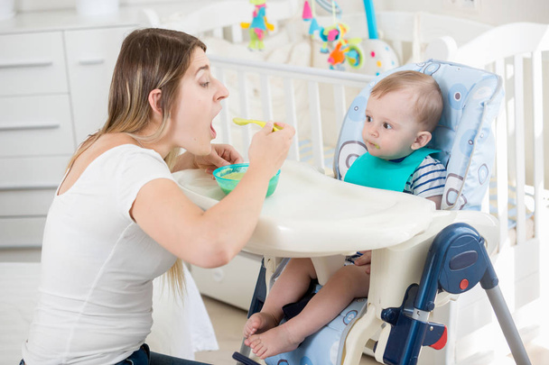 Sorrindo mulher alimentando seu bebê menino em cadeira alta na sala de estar
 - Foto, Imagem