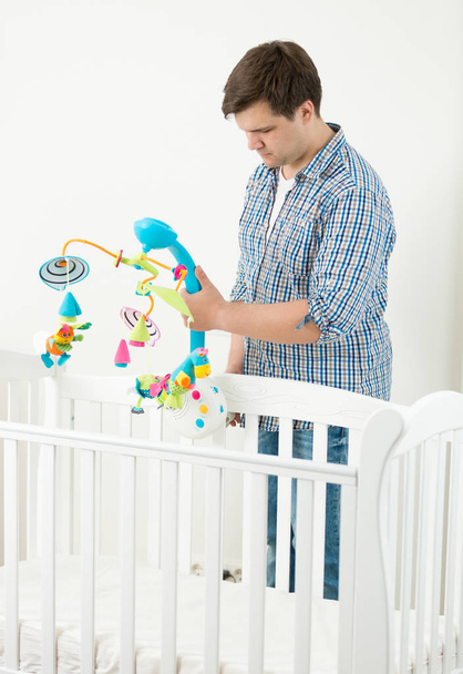 Joven hombre adjuntando carrusel de juguete en la cuna de su bebé
 - Foto, Imagen