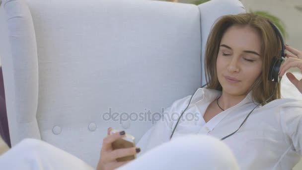Portrait of young caucasian woman in casual shirt listening music in headphones while relaxing on the chair in home. - Πλάνα, βίντεο