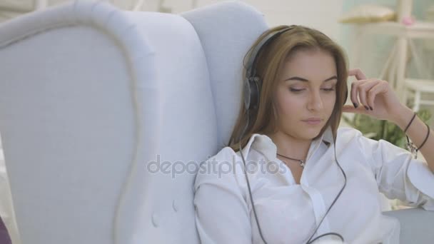 Young woman in casual shirt listening music in headphones while relaxing on the chair in home. Close up. - Video