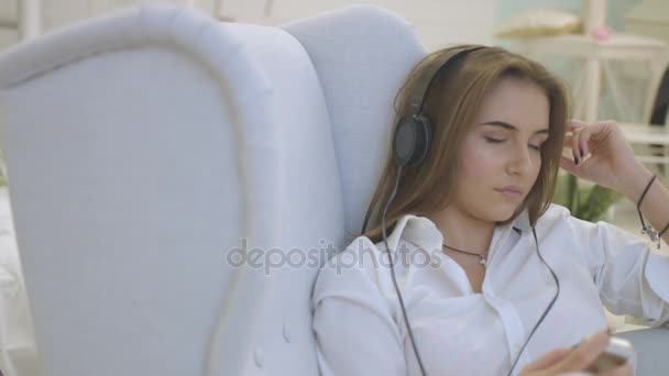 Young attractive woman in casual shirt listening music in headphones while relaxing on the chair in home. Close up. - Video