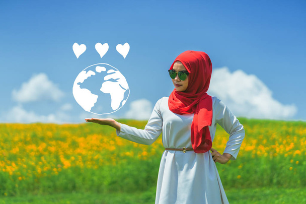 glücklich junge muslimische Frau Hand zeigt Liebe Erde über verschwommenem grünen Feld mit gelben Kosmos Blumen unter blauem bewölkten Himmel background.love earth Konzept. - Foto, Bild