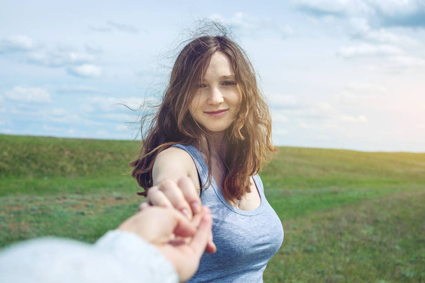 Follow me, Attractive brunette girl holding the hand of the leads in a clean green field, steppe with clouds - Photo, Image