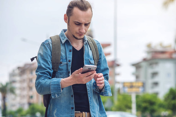 Mann mit Rucksack blickt auf sein Smartphone im Hintergrund der Stadt. - Foto, Bild
