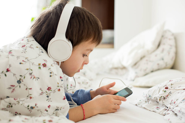 Little boy in bed, playing on phone, listening music on headphon - Foto, afbeelding