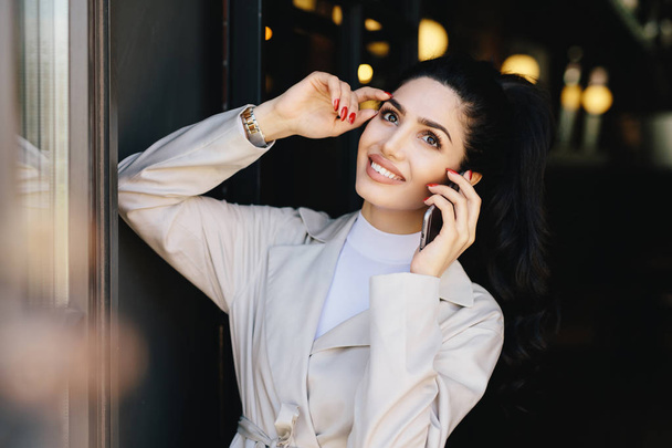 Horizontal portrait of beautiful lady with dark hair and eyes, make-up and lips smiling broadly holding hand on head while speaking over smartphone with her husband. Pretty woman in elegant clothes - Photo, Image