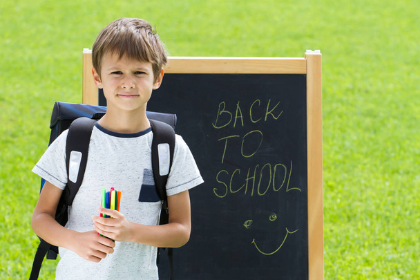 Little schoolboy with pens and backpack against the blackboard. Education, back to school concept - Foto, Imagem