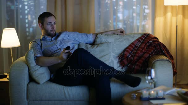 Ve středu večer letitý muž sedí na gauči, sledování televize, mění kanály s dálkovým ovládáním. Na pozadí Windows zobrazit velké město s mrakodrapy. - Záběry, video