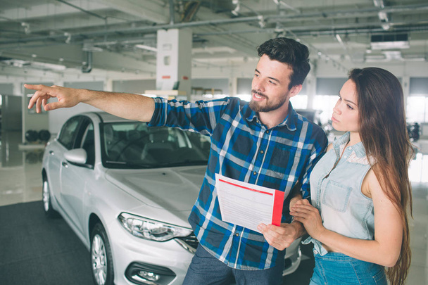 Happy couple chooses to buy a car in showroom. Beautiful young man and women in the auto show, choosing a car. Beautiful husband and wife standing at the car dealership and making their decision - Fotó, kép